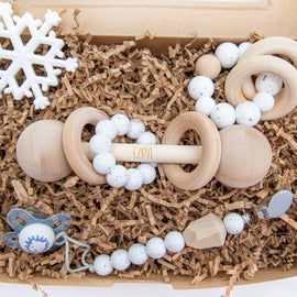 All-white newborn gift box with wooden rattle, pacifier clip, silicone snowflake teether, and round sensory teething ring displayed in kraft packaging