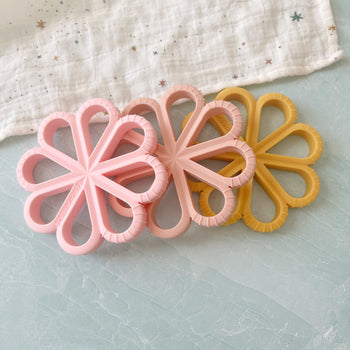 Flat lay of three flower-shaped silicone baby teethers in soft pink and mustard on pastel surface — Bead Sprout